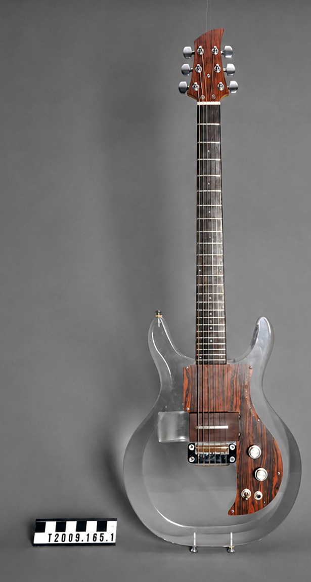 A clear acrylic-bodied electric guitar photographed against a neutral backdrop with a museum scale and accession number